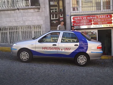Halıseren & UNY firma aracı (Fiat Palio) araç giydirme - gümüş/mavi kurumsal folyo kaplama, yan görünüm.
