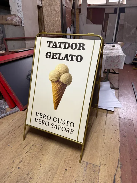 Tatdor Gelato için dondurma baskılı altın rengi çerçeveli ayaklı A pano, atölye zemininde çapraz görünüm.