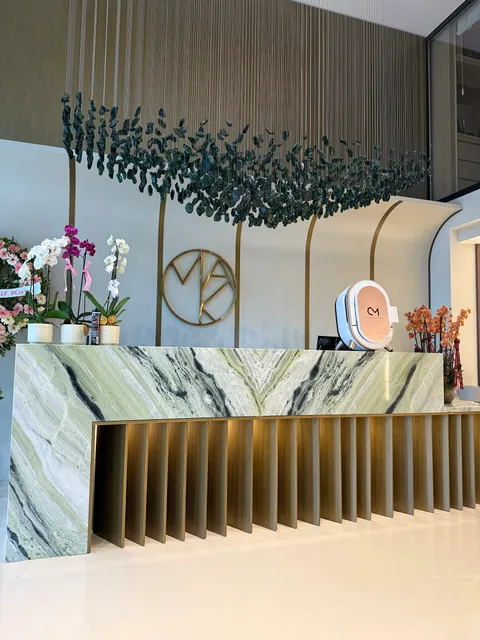 Altın renkli daire içinde MAK monogramlı metal duvar logosu, yeşil mermer yüzeyli ve pirinç çıtalı resepsiyon bankosunun arkasında.