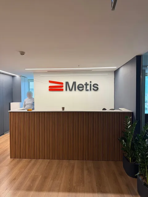 Metis için kırmızı simge ve antrasit harflerden oluşan kabartma logo, ahşap çıtalı resepsiyon bankosunun arkasındaki beyaz duvarda.