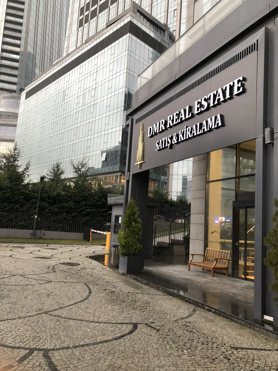 DMR Real Estate Satış & Kiralama için bina girişi üzerinde koyu panoda beyaz kabartma harfler, cam kuleler önünde gündüz görünüm.