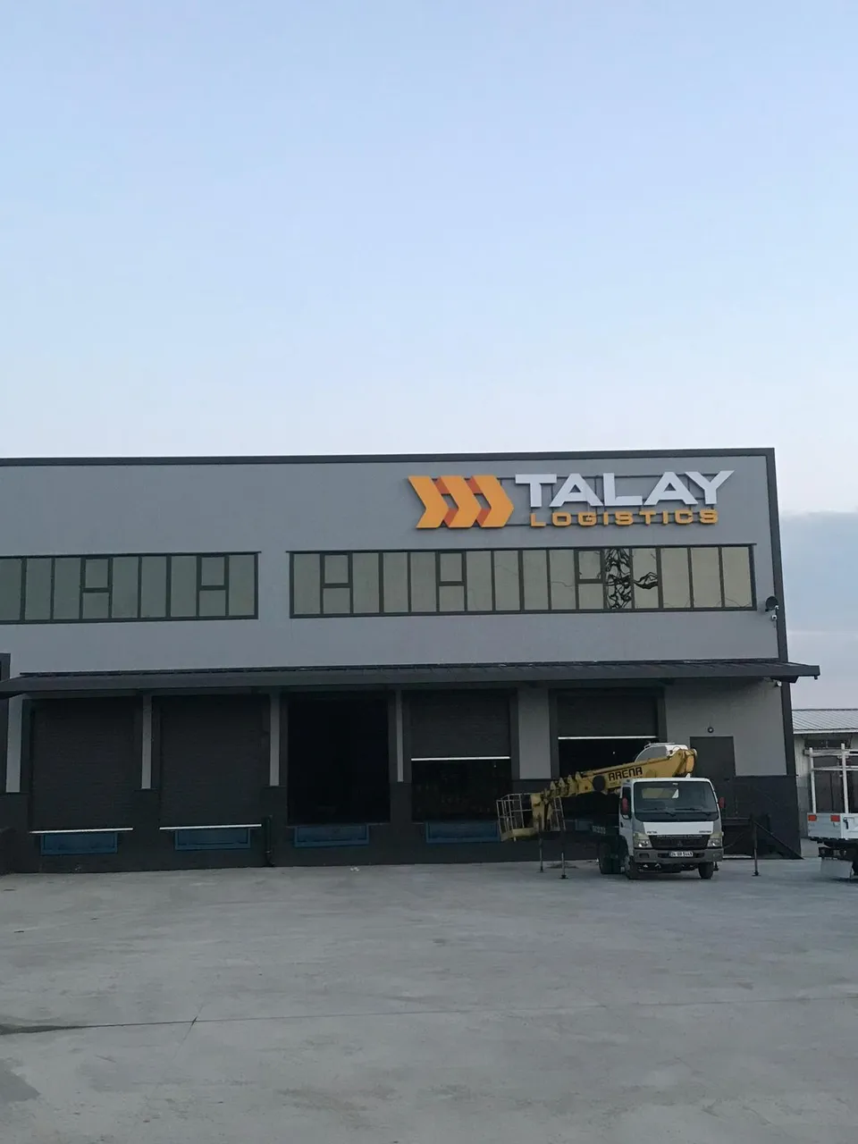Talay Logistics fabrika cephesinde sarı-gri tabela ve üretim sahası, geniş açı.