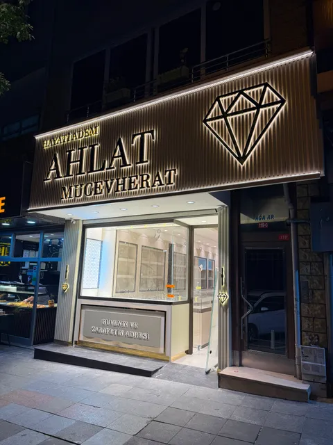 Hayati Adem Ahlat Mucevherat yazılı mağaza cephesinde dikey lamelli pano üzerinde arkadan ışıklı kabartma harfler ve elmas logo, gece, hafif yandan görünüm.