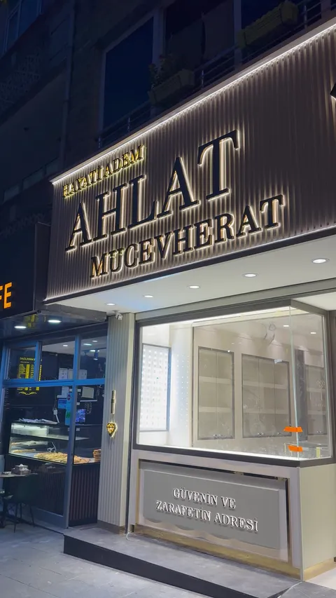 Hayati Adem Ahlat Mucevherat yazılı mağaza cephesinde dikey lamelli pano üzerinde arkadan ışıklı siyah büyük harfler ve altın renkli küçük harfler, gece yandan yakın görünüm.