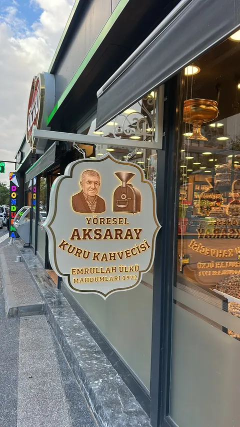 Yöresel Aksaray Kuru Kahvecisi için süslü beyaz konsoldan zincirle asılı, kıvrımlı kenarlı krem renkli fener tabela, altın çerçeve ve arkadan ışıklı kahverengi yazılarla akşamüstü yandan görünüm.