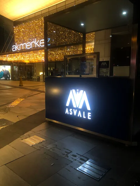 Asvale logolu lacivert banko üzerinde beyaz ışıklı logo, gece alışveriş merkezi girişinde yandan dikey görünüm.