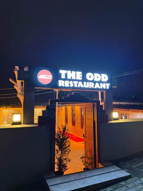 The Odd Restaurant girişinde beyaz ışıklı harfler ve kırmızı yuvarlak logolu siyah kutu tabela, gece görünümü.