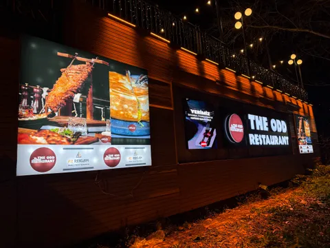 The Odd Restaurant için ahşap cephede beyaz ışıklı kutu harfli tabela, yemek baskılı ışıklı panolar ve LED ekran, gece görünümü.