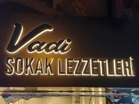 Vadi Sokak Lezzetleri yazılı siyah panelde arkadan sıcak beyaz ışıklı harfler ve altın aynalı yansımalar, gece karşıdan yakın görünüm.