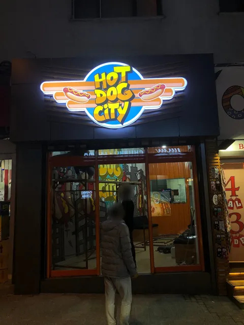 Hot Dog City için mavi, sarı ve turuncu kanatlı ışıklı logo tabelası, koyu panelli cephede gece görünümü.