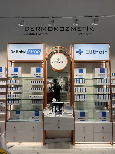 Beyaz duvarda siyah DERMOKOZMETİK harfleri, altında Dr.Balwi SHOP ve Elithair ışıklı panoları ile ortada Medicamine yuvarlak tabelası olan bakır çerçeveli mağaza rafları.