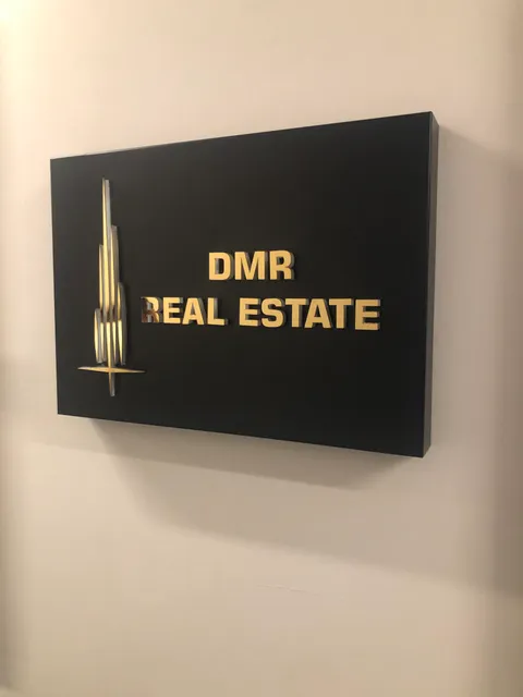 DMR Real Estate için siyah panoda altın aynalı kabartma harfler ve kule biçimli logo, krem duvarda çapraz görünüm.
