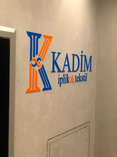 Kadim iplik & tekstil yazılı gri sıvalı duvarda mavi ve turuncu kabartma K logosu ve harfler, iç mekân yandan görünüm.