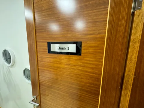 Klinik 2 yazılı kapı isimliği, siyah akrilik zemin üzerinde gümüş renkli fırçalı görünümlü plaka, ahşap kaplama kapıda yakın görünüm.