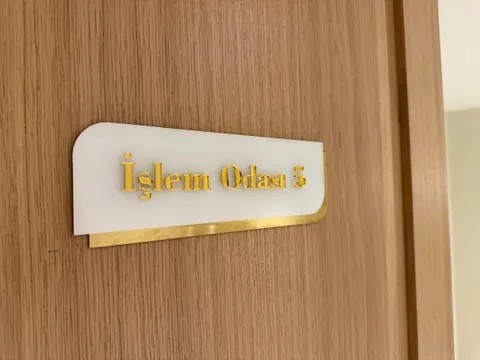 Ahşap kapıda beyaz levha üzerinde parlak altın harflerle İşlem Odası 5 yazan, altın fırçalı metal şeritli kapı isimliği.