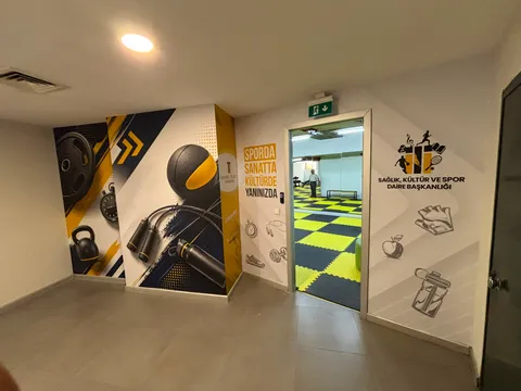 İstanbul Ticaret Üniversitesi spor alanı girişinde spor ekipmanı temalı sarı-lacivert duvar baskıları ve Sağlık, Kültür ve Spor Daire Başkanlığı yazılı duvar logosu, geniş görünüm.