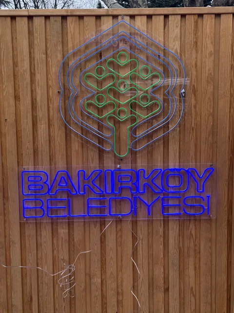 Ahşap kaplama yüzeyde şeffaf pleksi üzerine mavi ve yeşil neon ışıklı Bakırköy Belediyesi logosu ve yazısı, gündüz önden görünüm.
