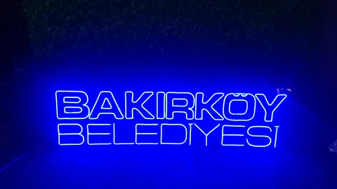 Bakırköy Belediyesi yazılı mavi neon harf tabelası, koyu yeşil yaprak duvarı önünde gece karşıdan görünüm.