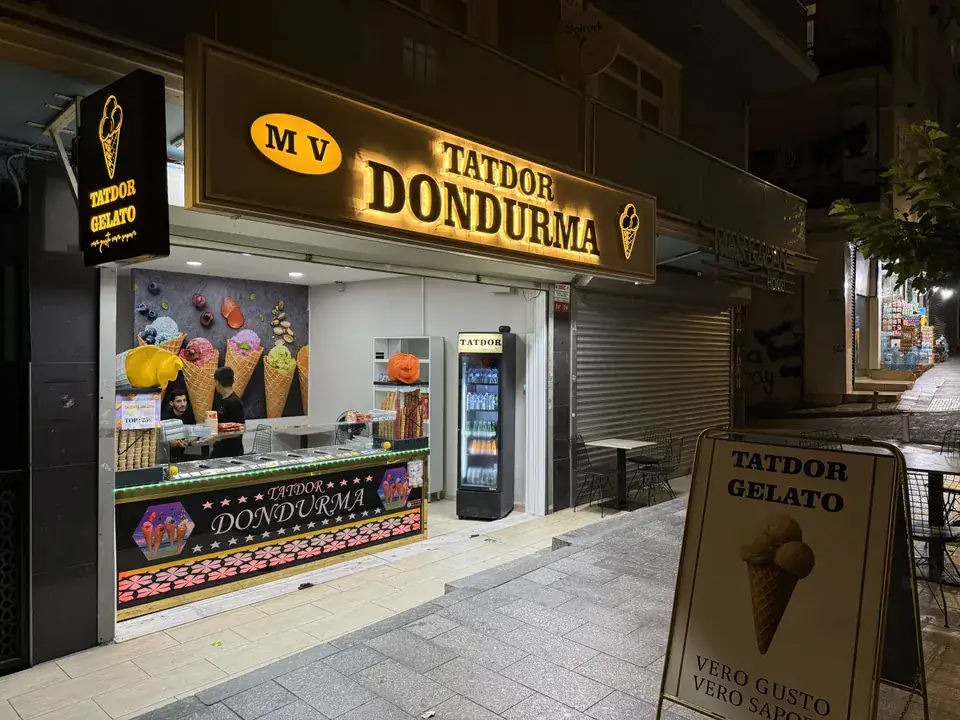 Tatdor MV Dondurma mağazasının altın ışıklı paslanmaz cephe tabelası, gece görünümü.
