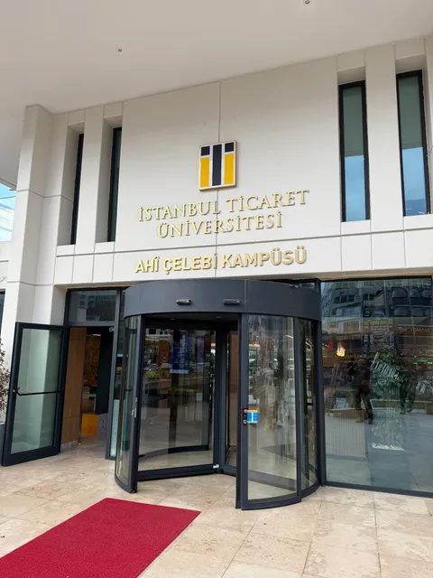 İstanbul Ticaret Üniversitesi Ahî Çelebi Kampüsü girişinde beyaz cephede altın renkli metal harfler ve sarı-siyah logo, döner kapı üstünde gündüz alttan görünüm.