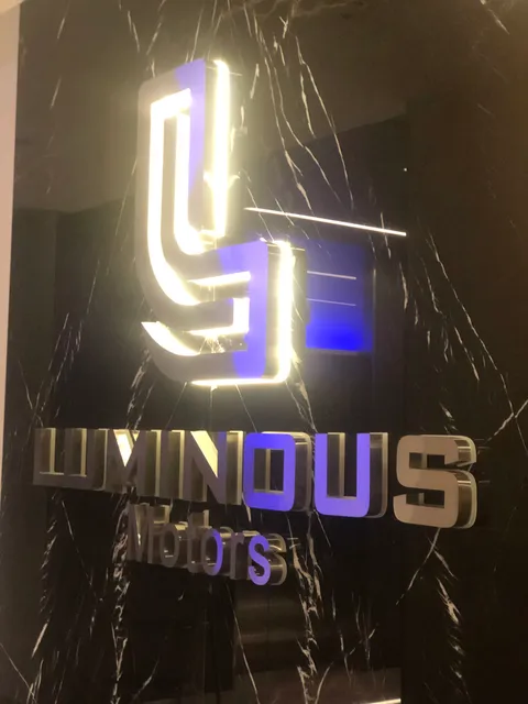 Luminous Motors için siyah mermer duvarda ışıklı paslanmaz logo ve harfler, mavi yansımalı iç mekân görünümü.