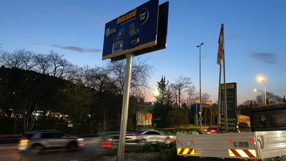 Yoğun trafikli kavşakta çift yüzlü yüksek reklam totemi, akşam görünümü.