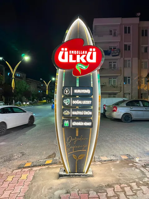 Emrullah Ülkü Kuruyemiş için ayçiçeği çekirdeği biçiminde ışıklı totem, kırmızı logo, dört ışıklı bilgi kutusu ve altın şeritler, gece önden görünüm.