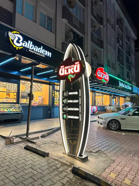 Emrullah Ülkü Kuruyemiş için ayçiçeği çekirdeği biçiminde ışıklı totem ve solda Balbadem Pastaneleri ışıklı cephe tabelası, gece yandan geniş görünüm.
